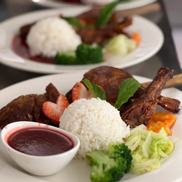 Two plates of Thai duck with rice, broccoli, cabbage, carrots, strawberries, and sauce.