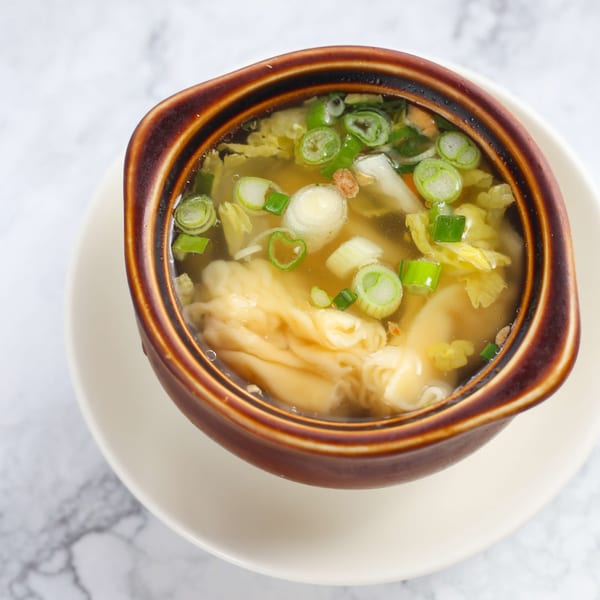 Wonton soup with scallions and vegetables in a brown bowl on a white plate.